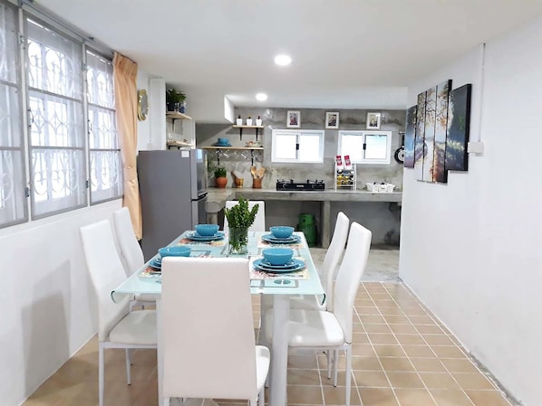 A dining area with a table set for four, white chairs, and a kitchen in the background with a refrigerator, stove, and various kitchen items.