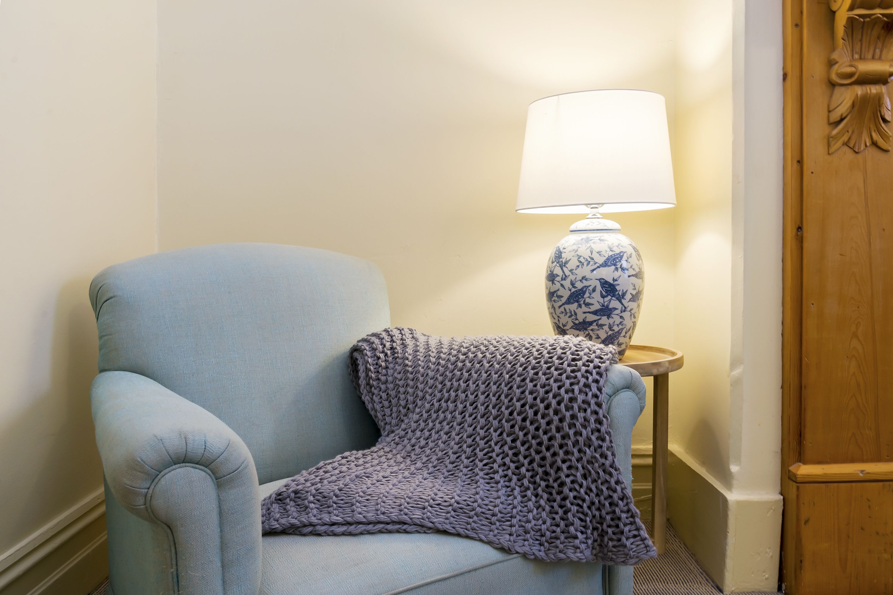 A light blue armchair with a knitted throw, a white lamp with a blue floral base, and a wooden door with a decorative handle.