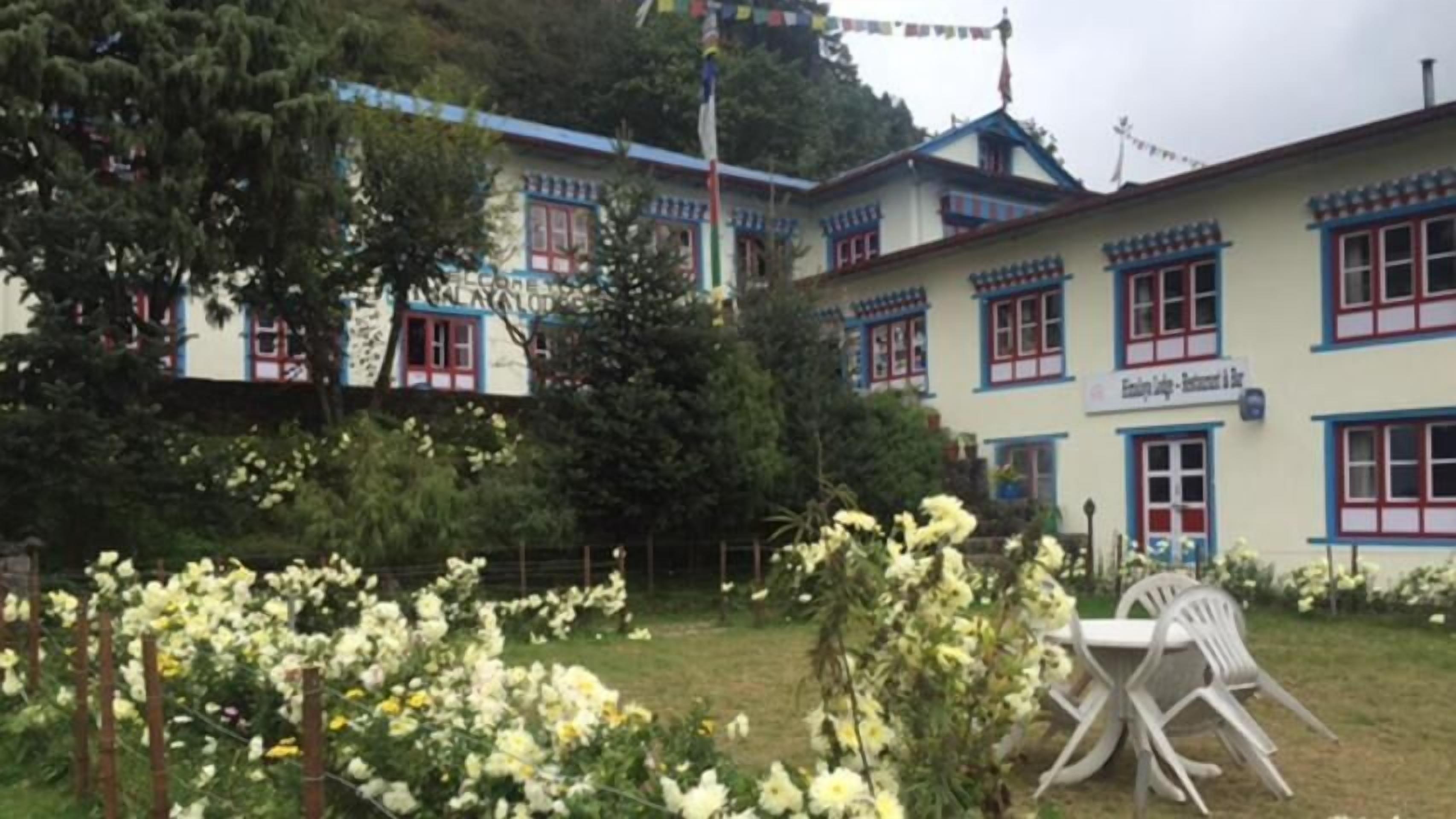 A building with multiple windows and a sign that reads "Hotel". There is a garden with flowers and a table with chairs in the foreground.