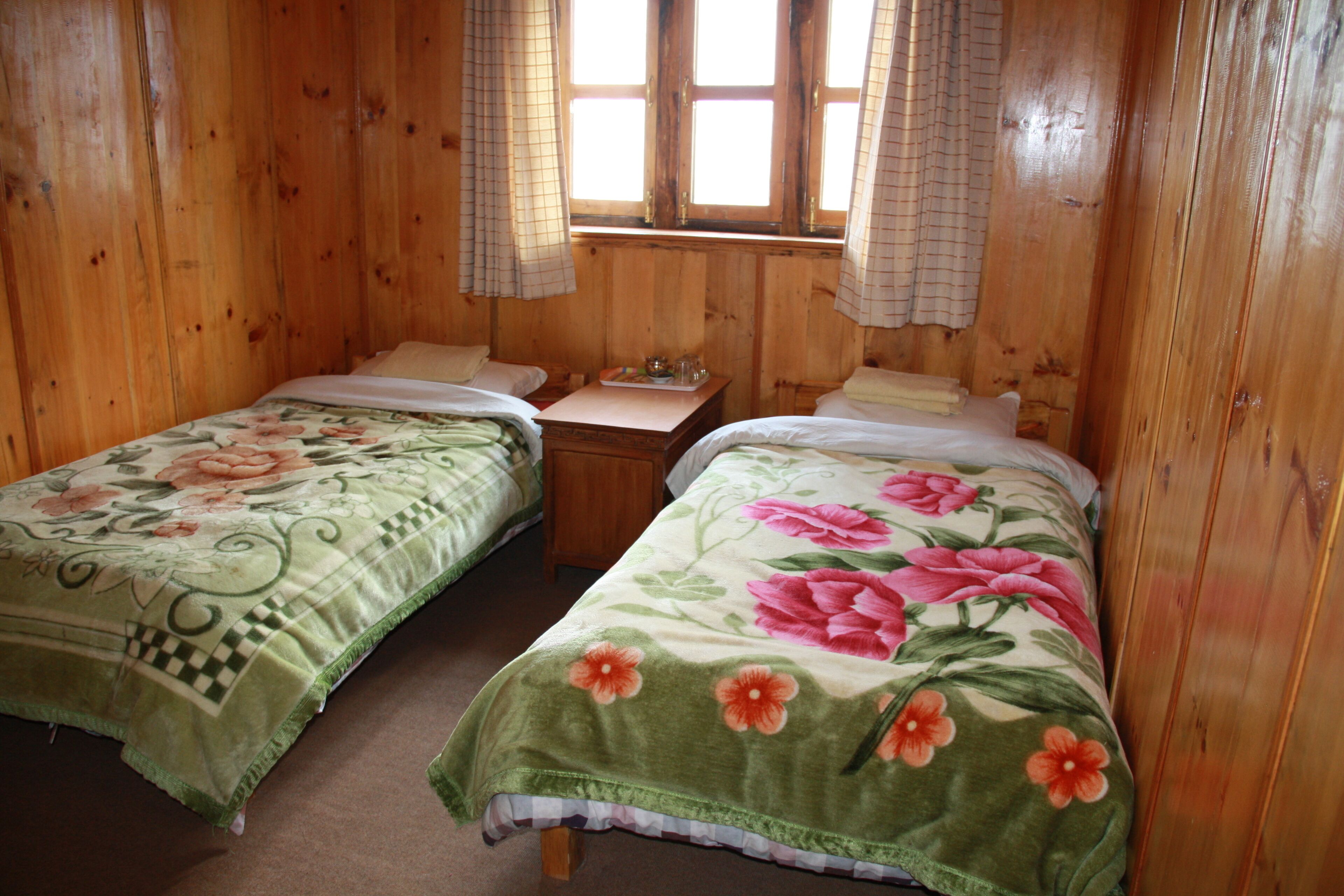 Ein Holzhütten-Zimmer mit zwei Betten, jeweils mit floraler Bettwäsche, einem kleinen Holztisch und einem Fenster mit Vorhängen.