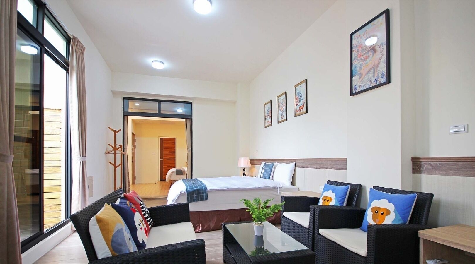 Une chambre d’hôtel avec un lit, deux canapés, une petite table et une plante en pot. Il y a une fenêtre avec des rideaux, une porte qui donne sur une autre pièce et un mur orné de tableaux encadrés.