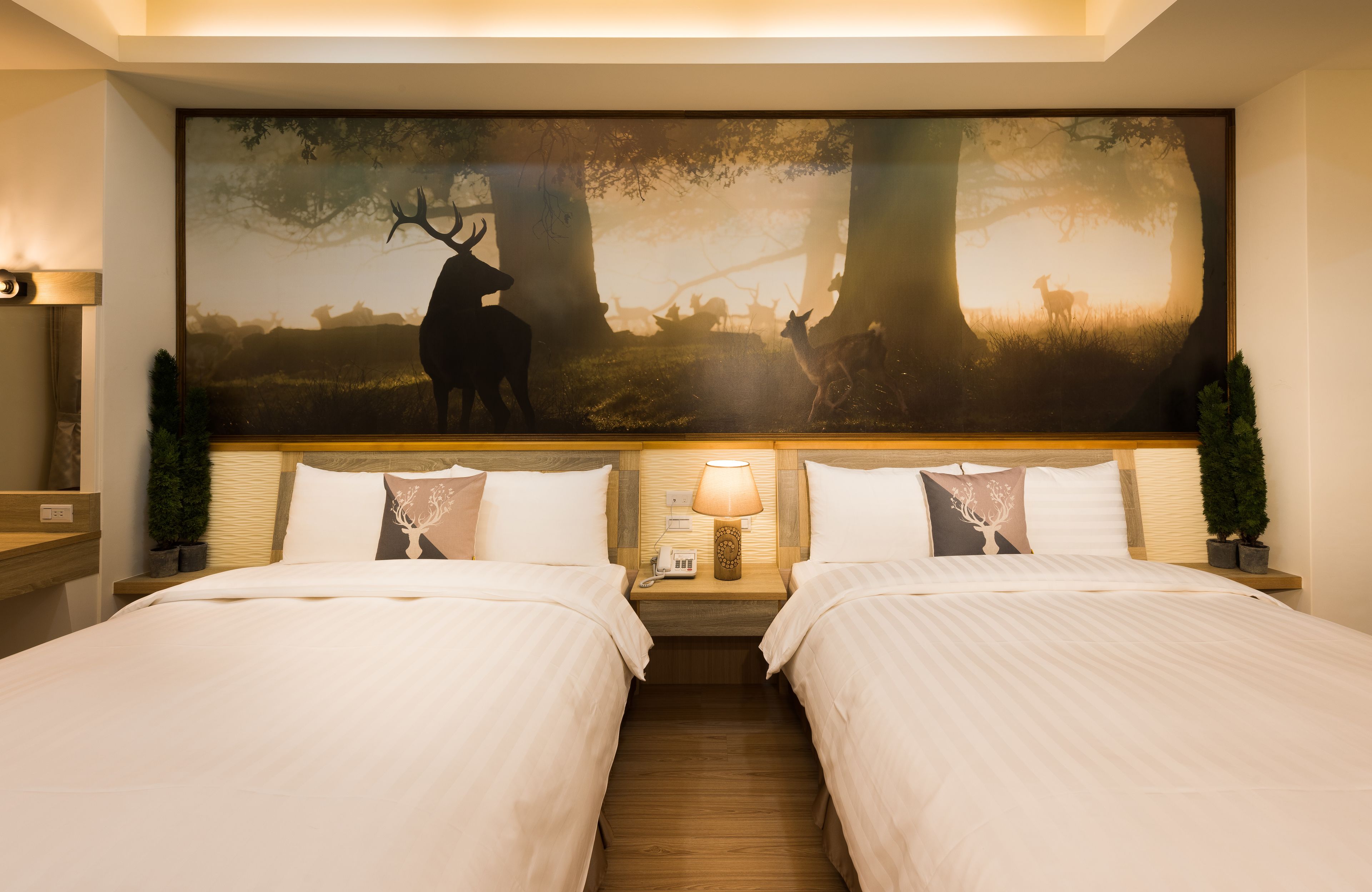 A hotel room with two beds, a large forest-themed mural on the wall, a nightstand with a lamp, and a small potted plant.