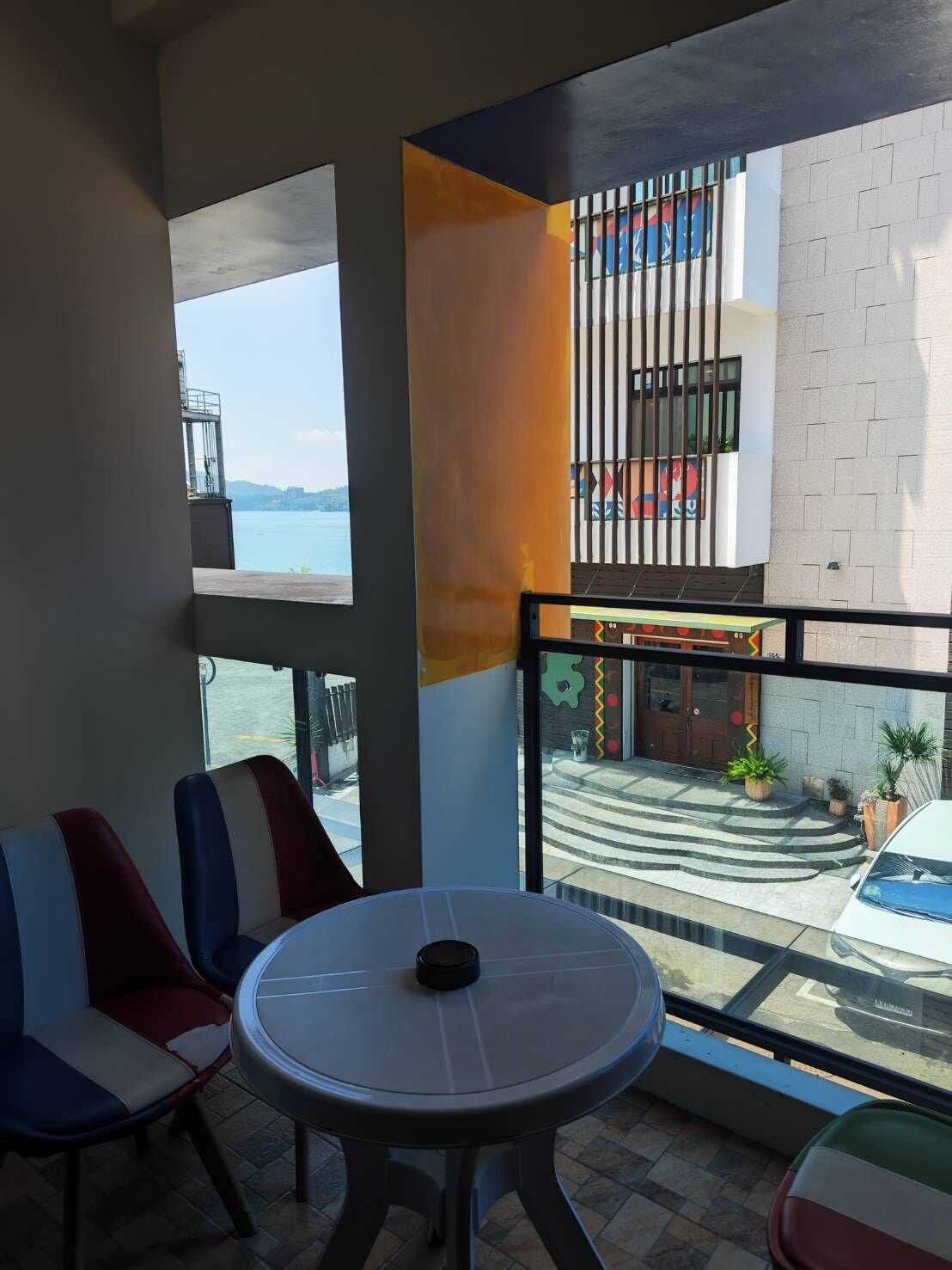 Un balcón con mesa y sillas, vista a un edificio con ilustraciones coloridas y una masa de agua al fondo.