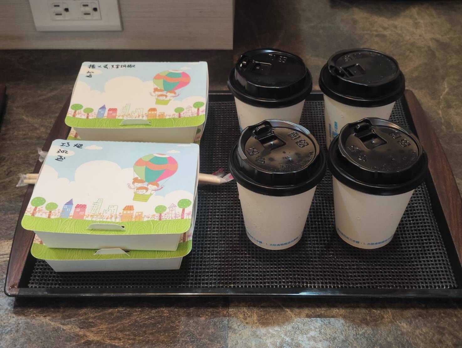 A tray with four disposable coffee cups and two insulated containers with lids, placed on a textured mat.