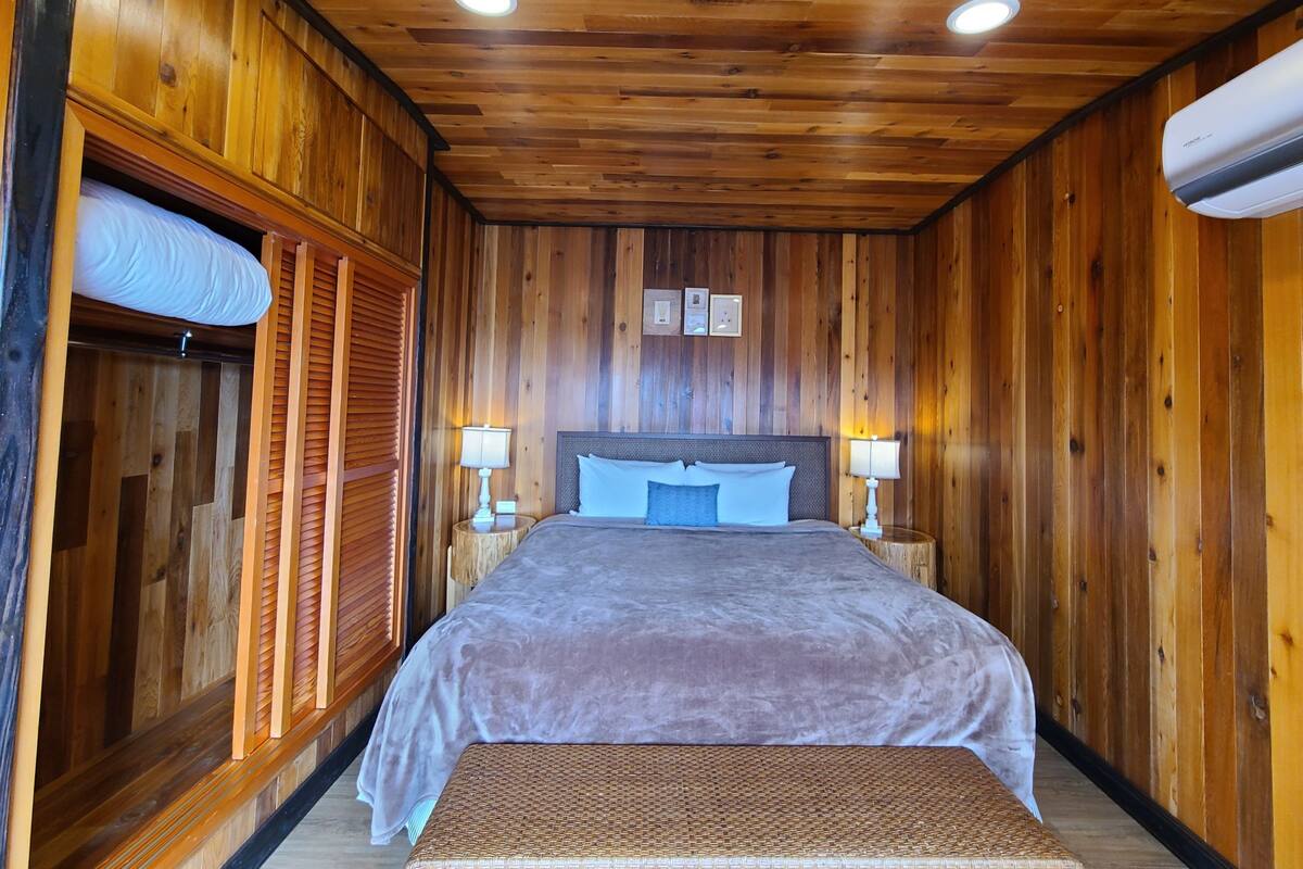 A wooden-paneled room with a bed, two bedside lamps, and a window with blinds.