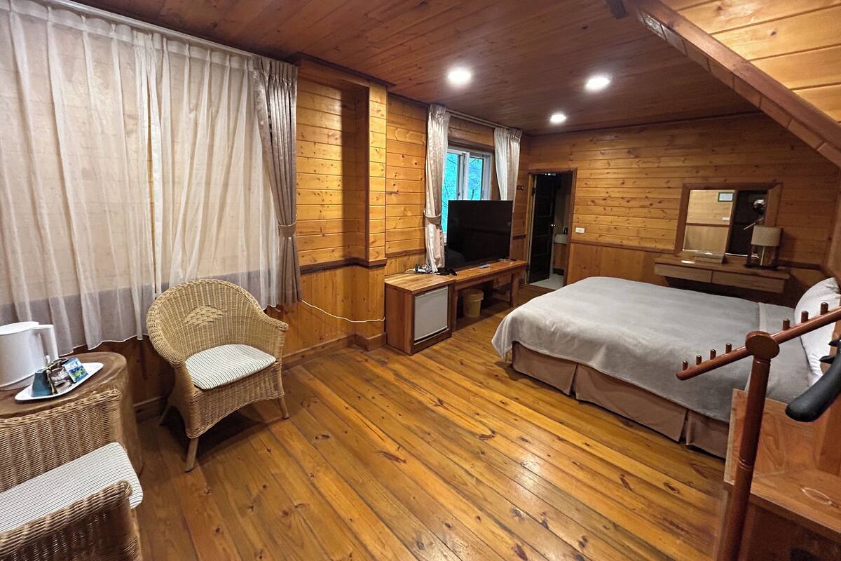 A wooden cabin room with a bed, a television, a small table, and wicker furniture.