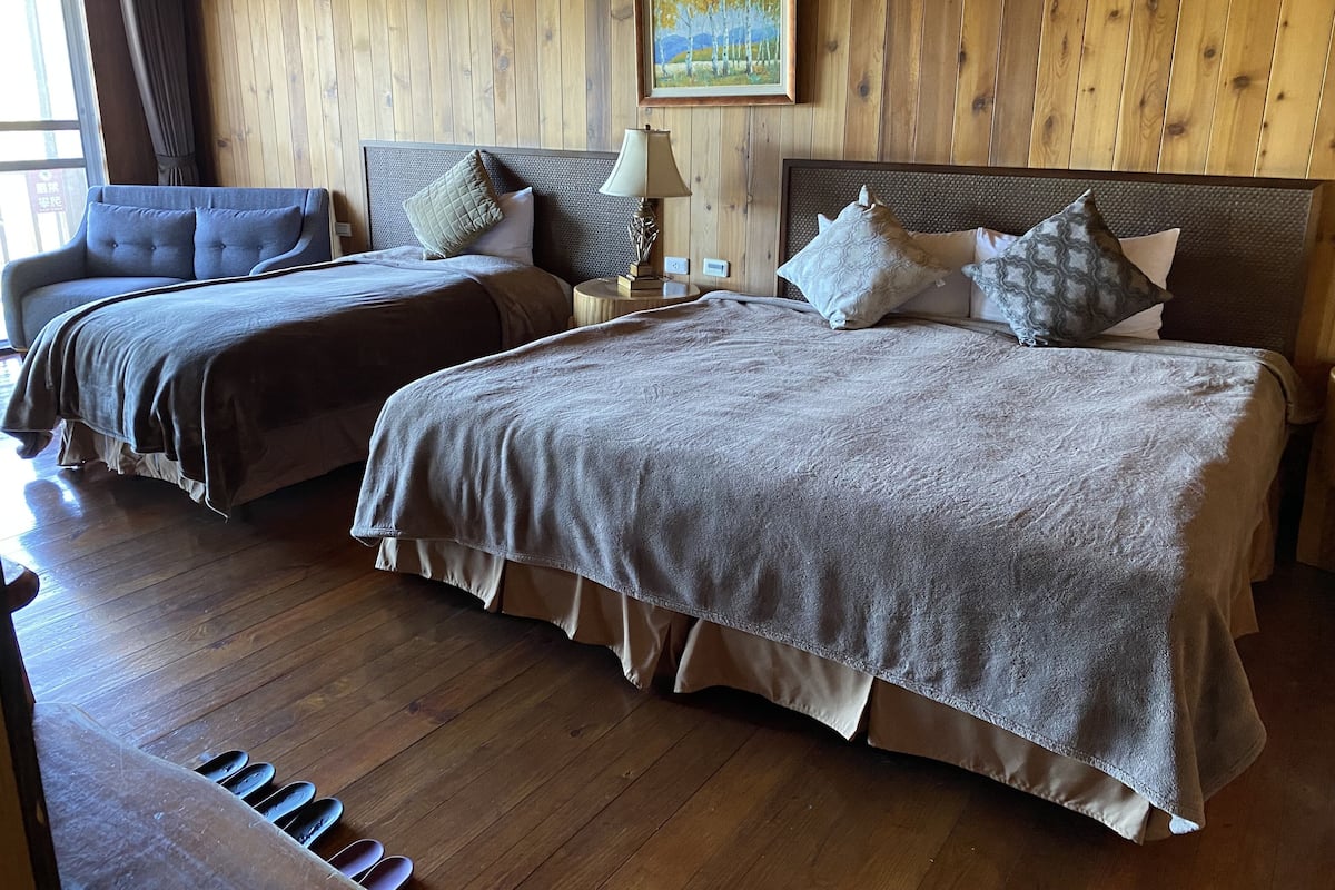 A room with two beds, wooden flooring, a sofa, a lamp, and a painting on the wall.