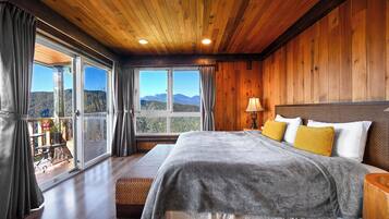 Quarto com painéis de madeira, uma cama grande, um banco e vista para montanhas pelas janelas.