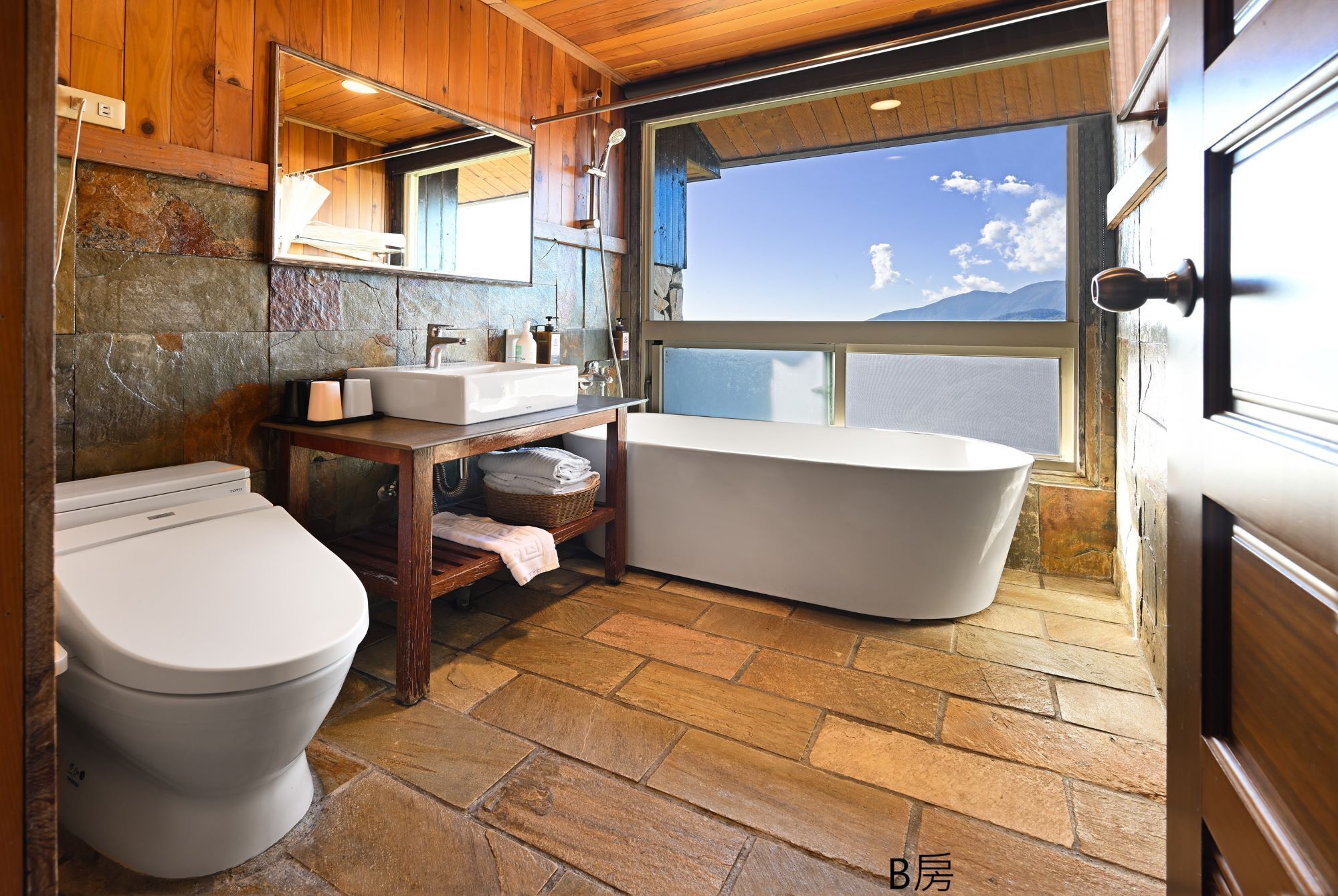 Un baño con pared de piedra, techo de madera, una bañera independiente, un inodoro y un lavabo con artículos de aseo.