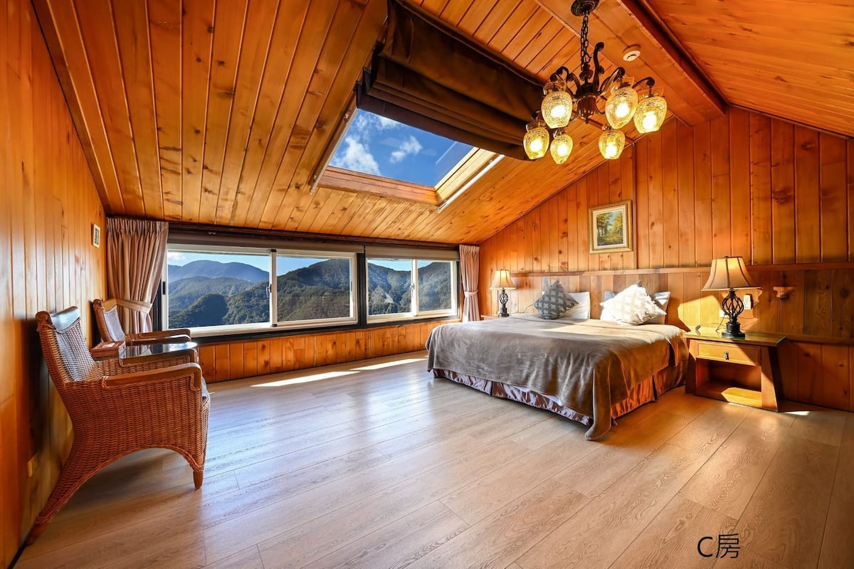 A cozy wooden cabin bedroom with a large bed, a wicker chair, a nightstand, and a view of mountains through the window.