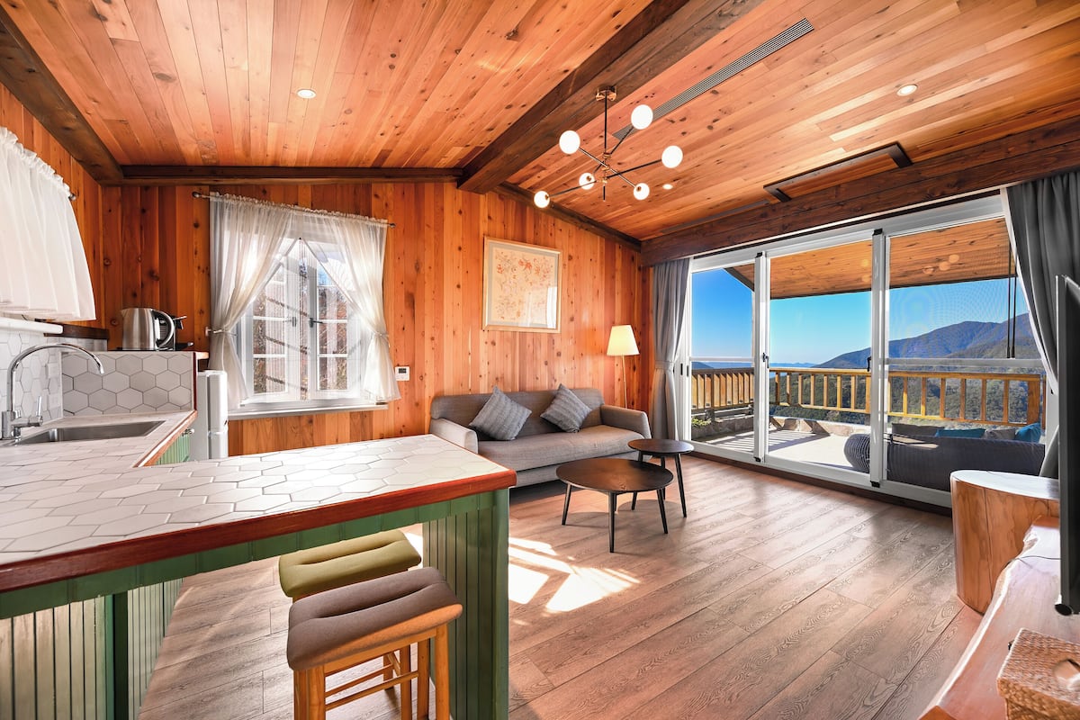 A wooden cabin interior with a kitchen, dining area, and a balcony with a mountain view.
