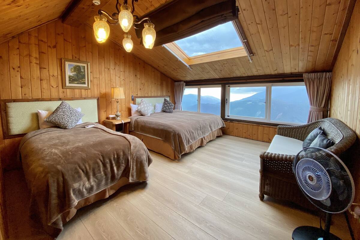 A cozy wooden cabin bedroom with two beds, a sitting area, and a large window offering a mountain view.