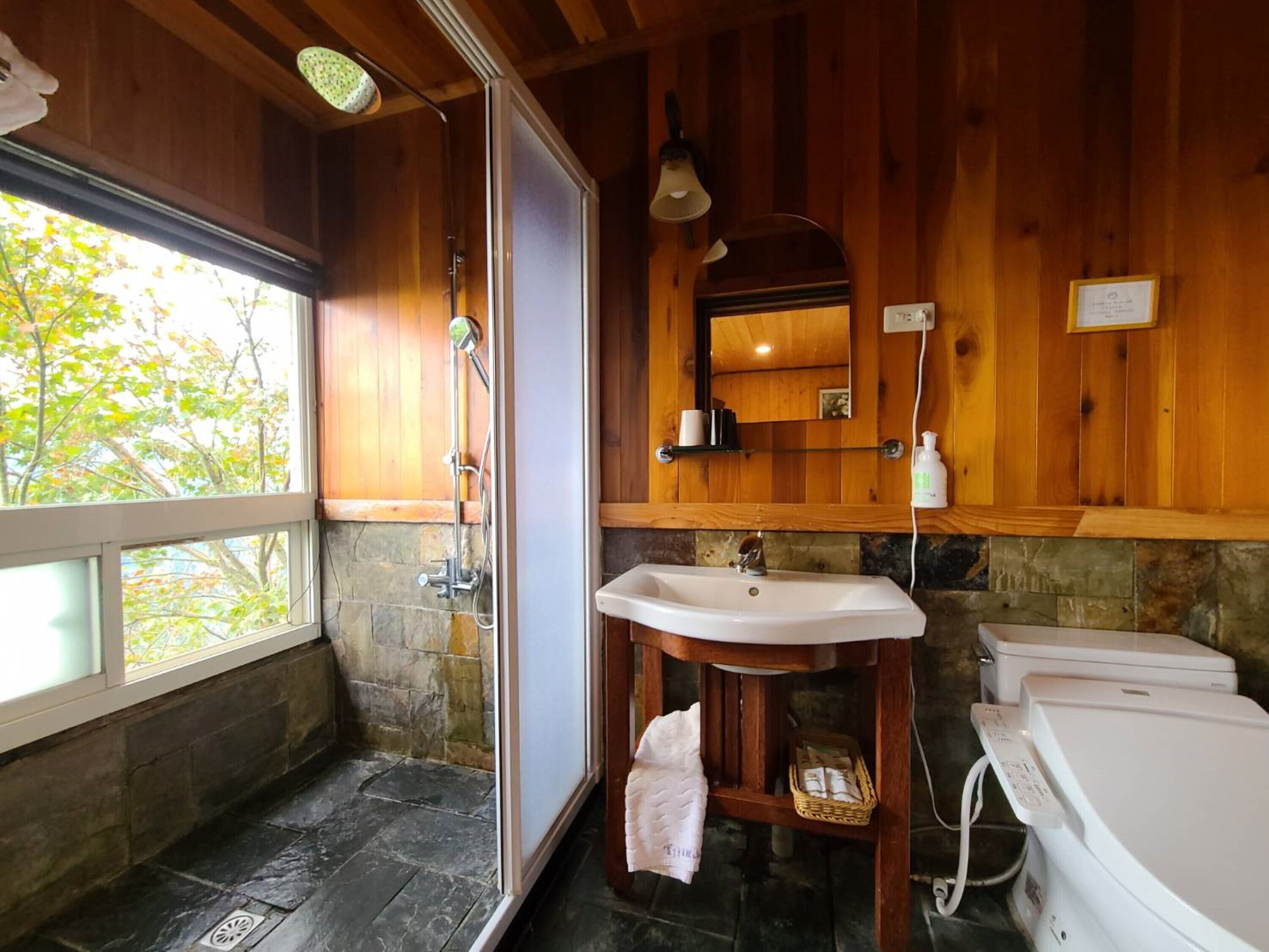 Une salle de bain avec un sol en pierre, des murs en bois, un lavabo blanc, un miroir, une douche et une baignoire.