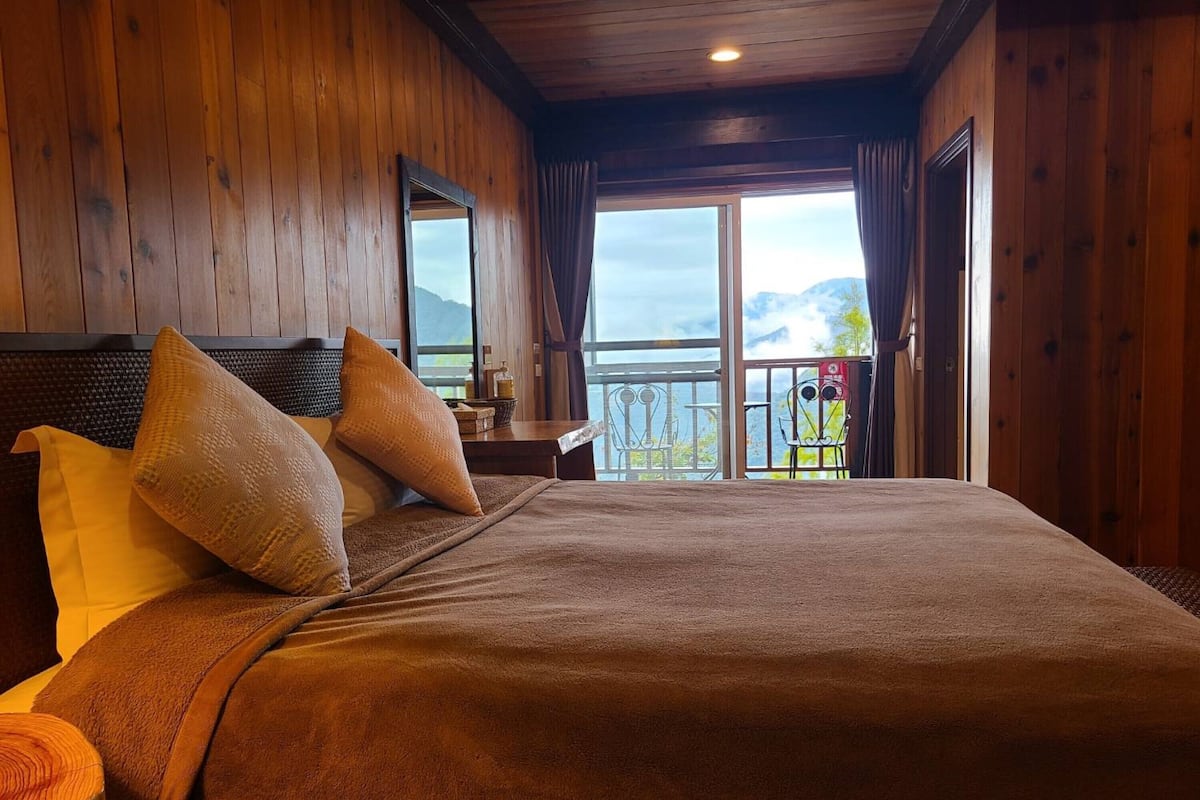 A wooden room with a large bed, two pillows, a nightstand, and a view of mountains through a balcony.
