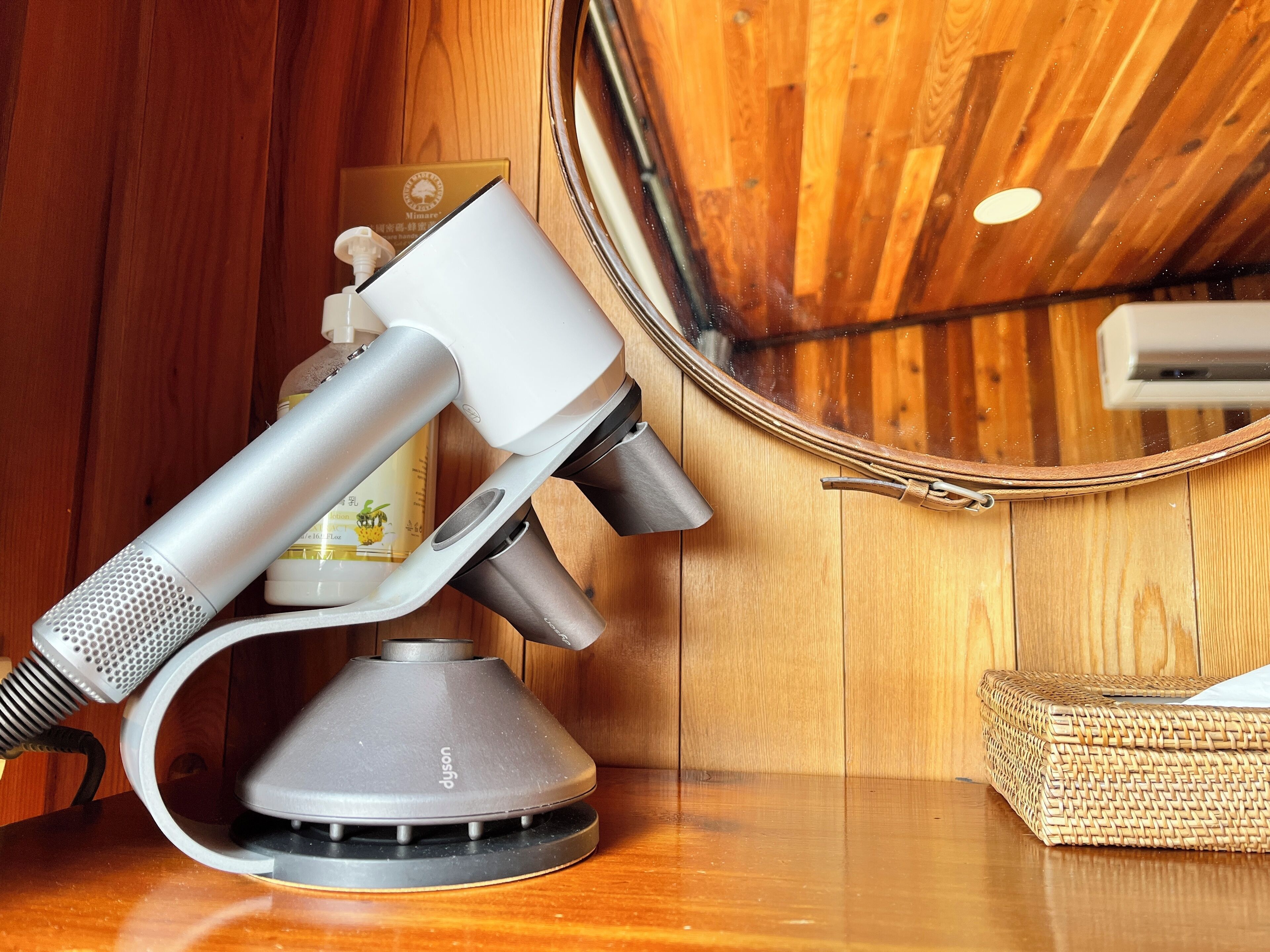 Um secador de cabelo Dyson e uma garrafa branca sobre uma mesa de madeira em um quarto com paredes de madeira e um espelho redondo.