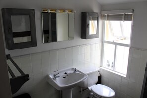 Et badeværelse med en hvid vask, toilet og et spejl. Der er et vindue med persienner og en bøjle til håndklæder.
