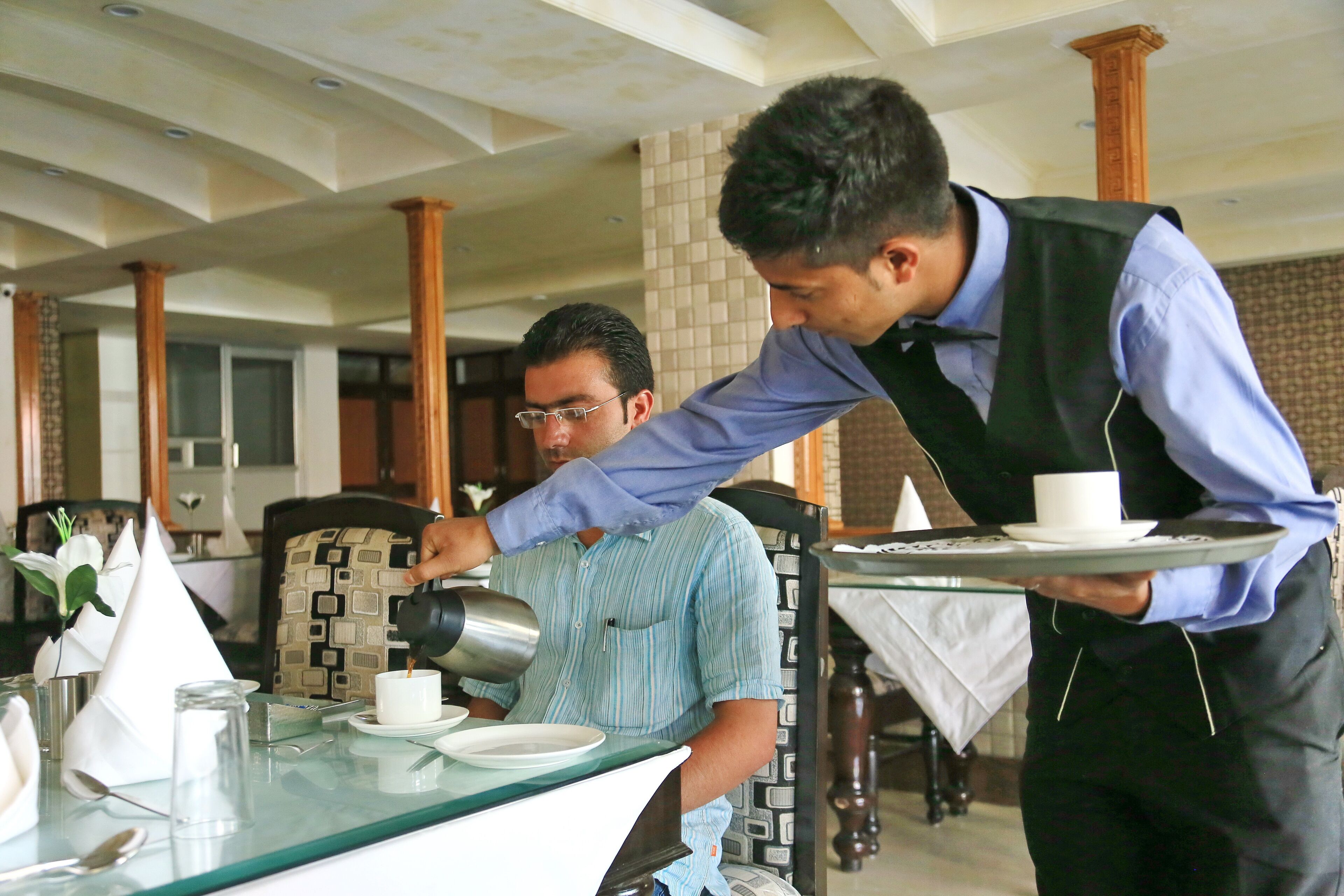 A waiter serving a cup to a seated guest in a restaurant with a modern interior, including patterned chairs and a glass table.