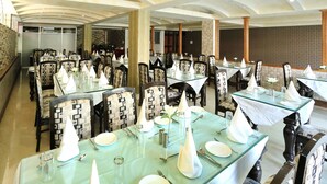 A restaurant with multiple tables set for dining, featuring patterned chairs and glass tables. - Crown Plaza Residency (Srinagar)
