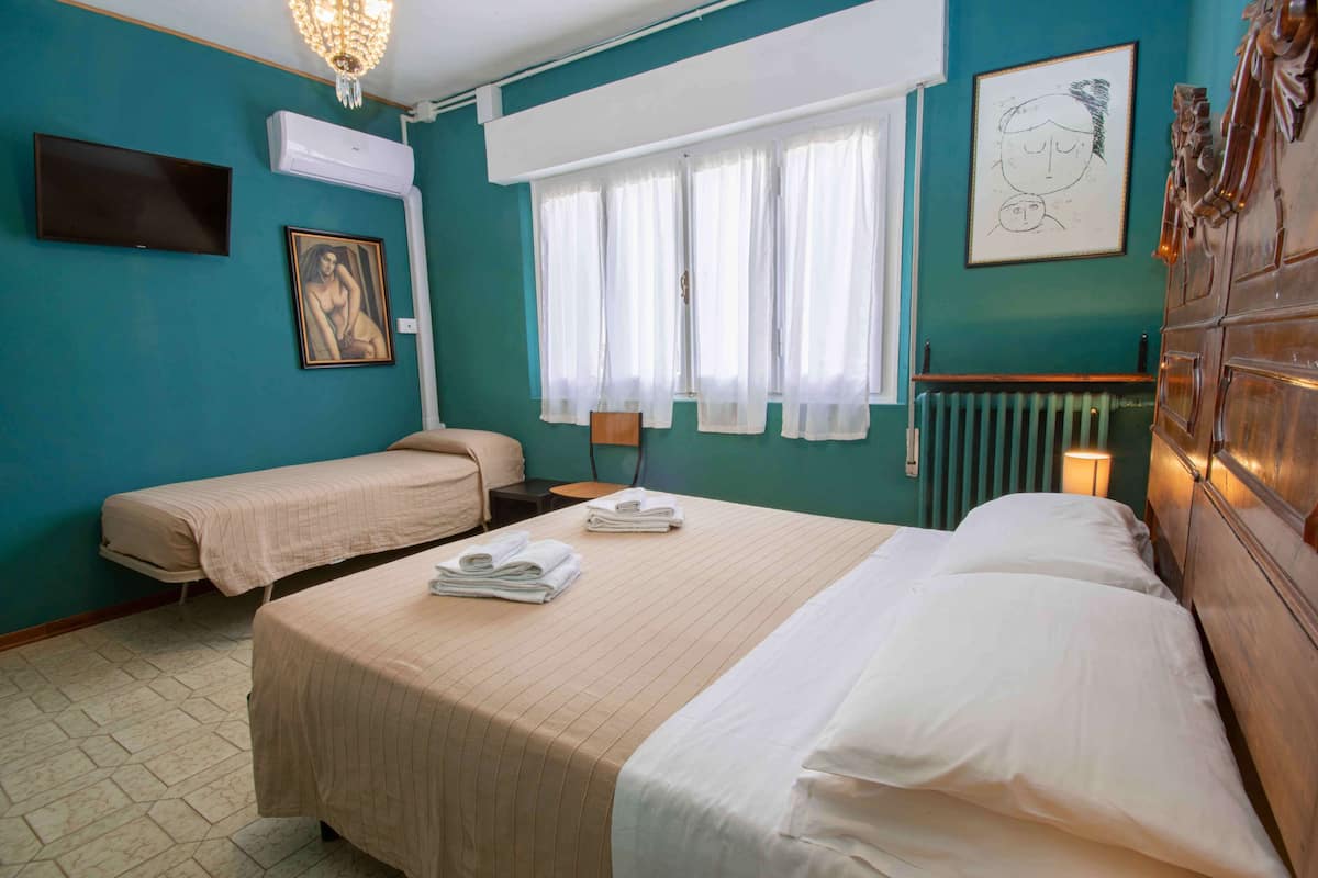 Habitación de hotel con dos camas, televisión, aire acondicionado, ventana con cortinas, radiador y un cuadro en la pared.