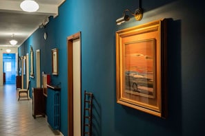 A hallway with blue walls, framed artwork, a wooden door, a chair, and a ladder. - Hotel La Villa (Cervia)