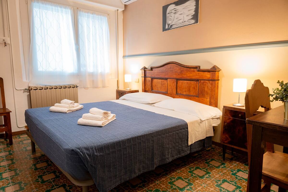 Una cama bien hecha con colcha azul y ropa de cama blanca, cabecera de madera, dos lámparas de noche, un radiador y baldosas de suelo con diseño.