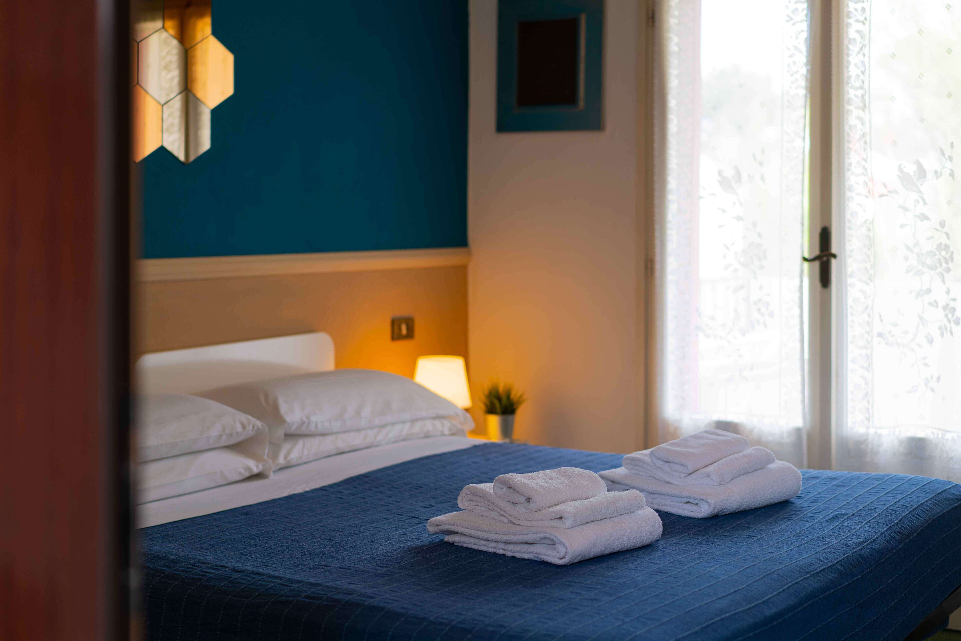 Ein ordentlich bezogenes Bett mit blauer Bettwäsche und weißen Kissen, ein kleiner Nachttisch mit einer Lampe sowie ein Fenster mit blumigem Vorhang.