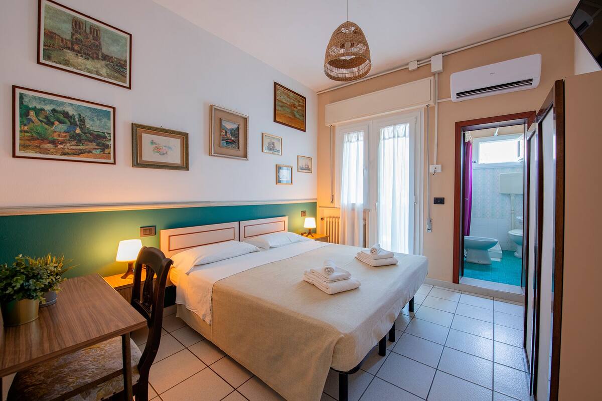 Habitación de hotel con cama, mesita de noche, lámpara y vistas a un espacio exterior.