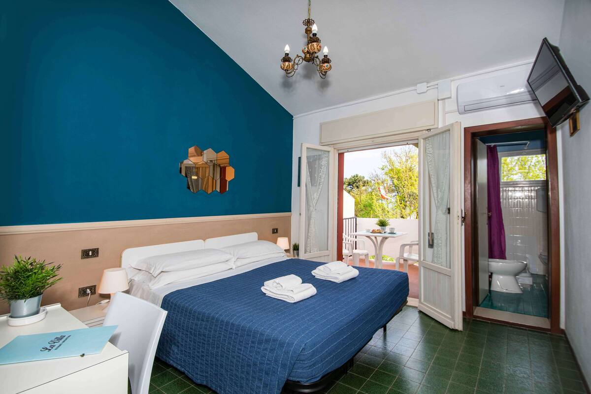 Habitación de hotel con una cama grande, un escritorio con una planta, una araña y vistas al exterior.