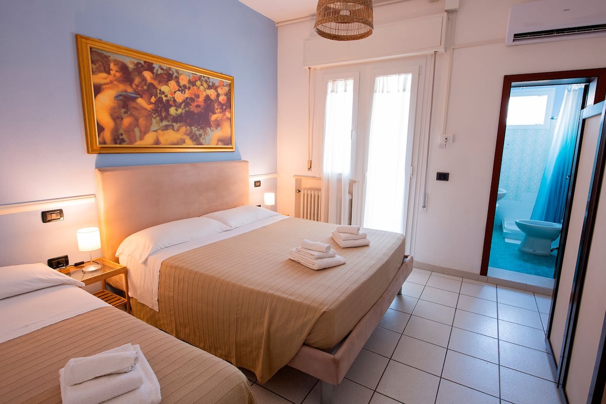 Habitación de hotel con dos camas, un cuadro grande, un baño con inodoro azul y una puerta que da a un balcón.