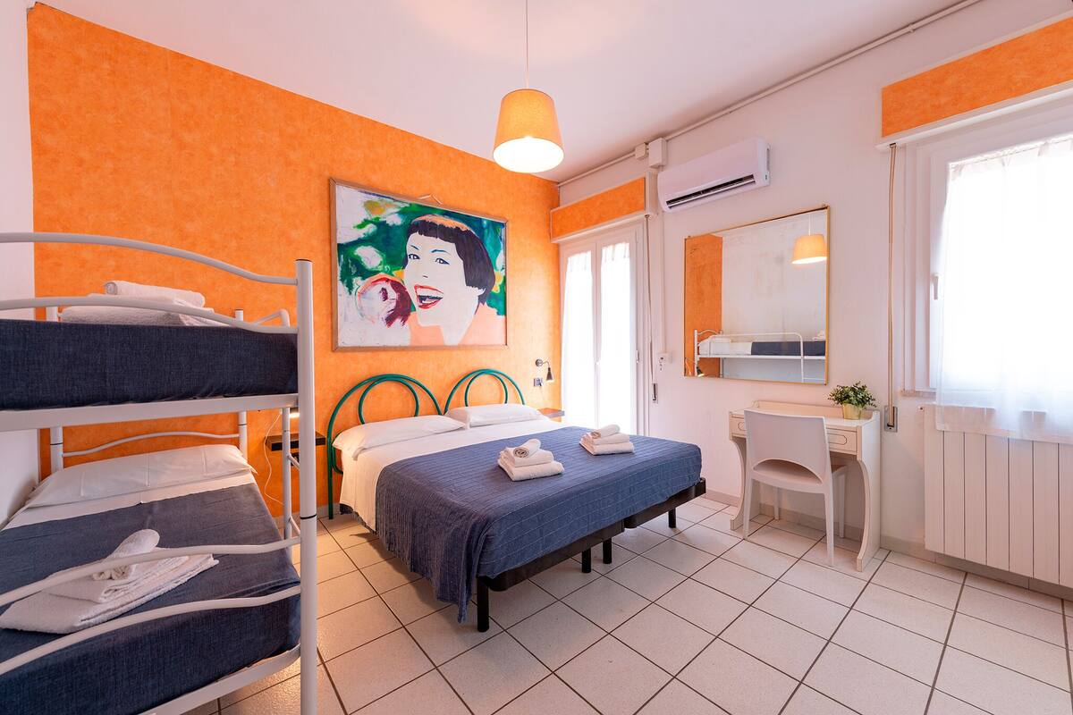 Habitación de hotel con literas, un escritorio, una silla, una cama con manta azul, un cuadro en la pared y un equipo de aire acondicionado.