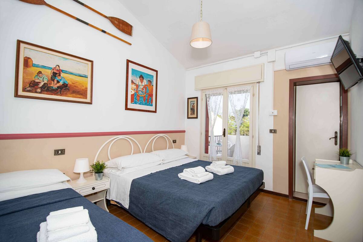Habitación de hotel con dos camas, un escritorio, una silla, un televisor y obras de arte en las paredes.