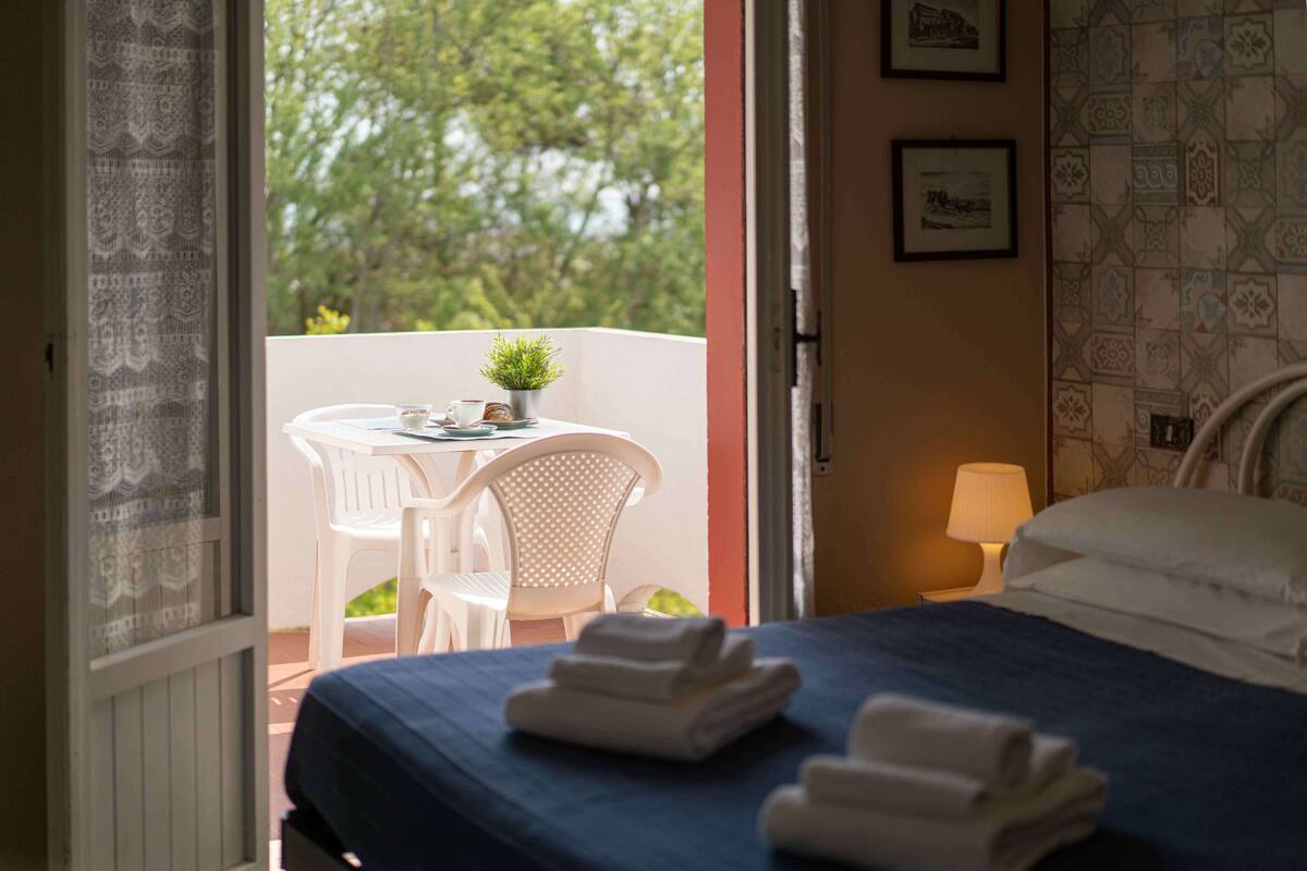 Habitación de hotel con cama, un pequeño balcón con mesa y sillas, y vista a árboles.