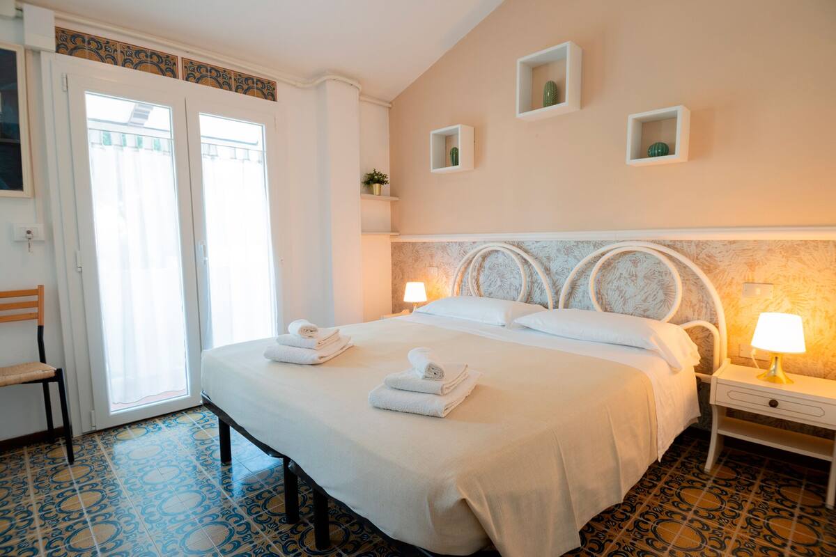 Habitación de hotel con una cama grande, dos mesitas de noche, una silla y una puerta que da a un balcón.