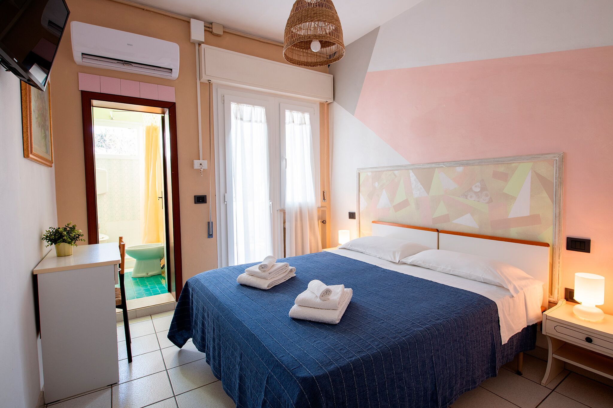A hotel room with a bed, a bedside table with a lamp, a small desk with a plant, and a bathroom visible through an open door.