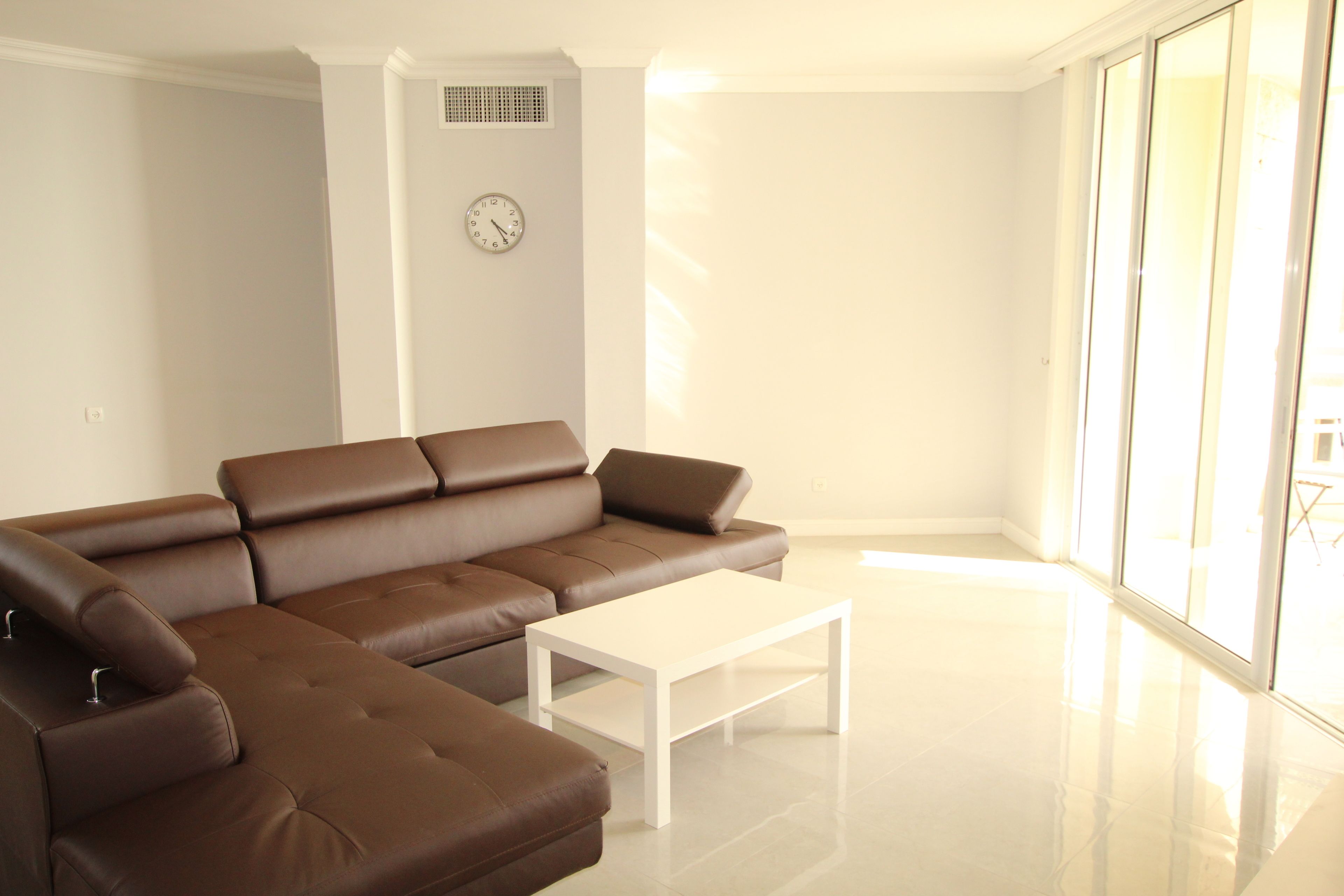 Un salón moderno con un sofá seccional marrón, una mesa de centro blanca y un amplio ventanal que proporciona luz natural.