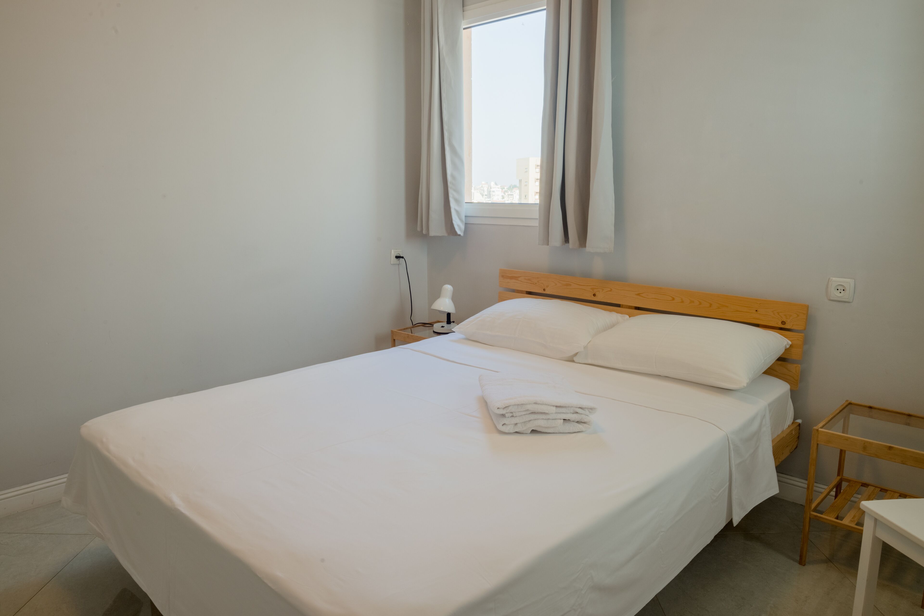 A neatly made bed with white linens and a folded towel, a wooden headboard, a bedside table, and a window with curtains.