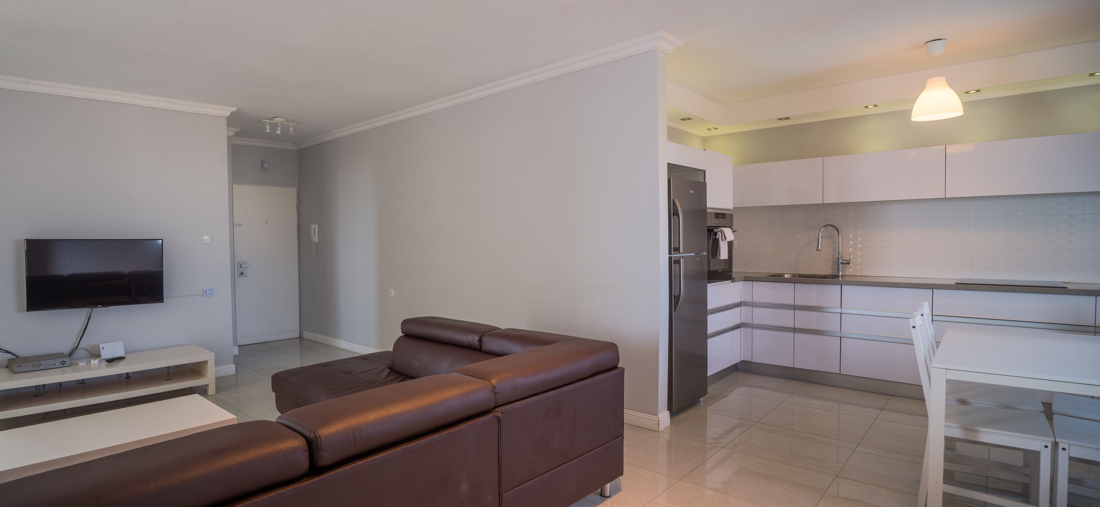 Een moderne woonkamer met een bruine leren bank, een flatscreen televisie en een klein tafeltje. De keuken heeft witte kasten en een roestvrijstalen koelkast.