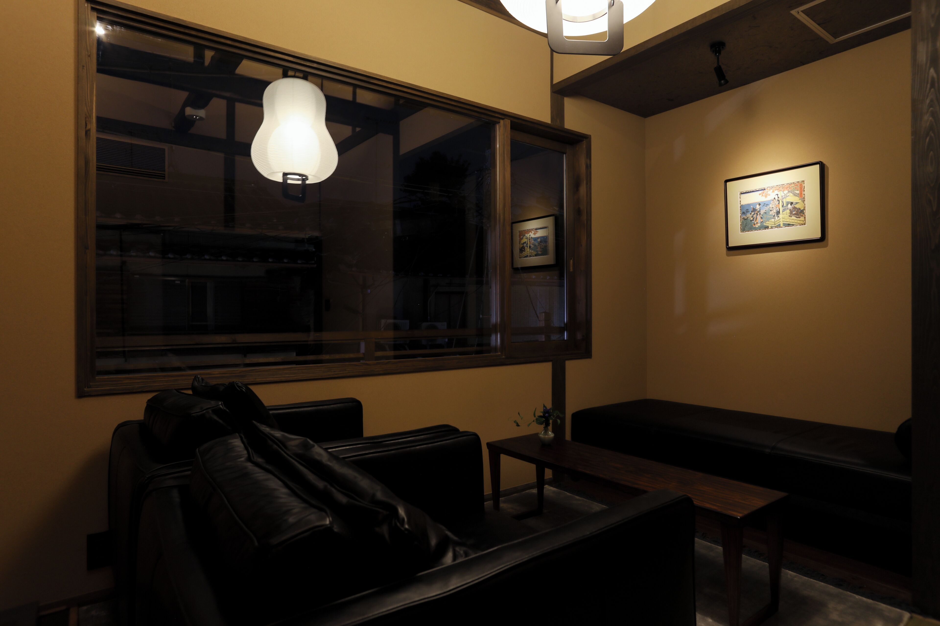 A dimly lit room with a large window, a bench, a table, and a framed picture on the wall.