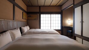 Una tradizionale stanza in stile giapponese con pavimento in tatami, soffitto in legno basso e porte scorrevoli shoji. Si notano due letti con biancheria da letto bianca e un piccolo tavolo con una sedia.
