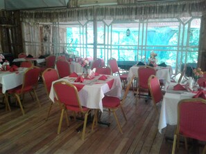 A dining area with multiple tables set for guests, featuring red chairs and white tablecloths. - Brooks Lodge (Gilgil)