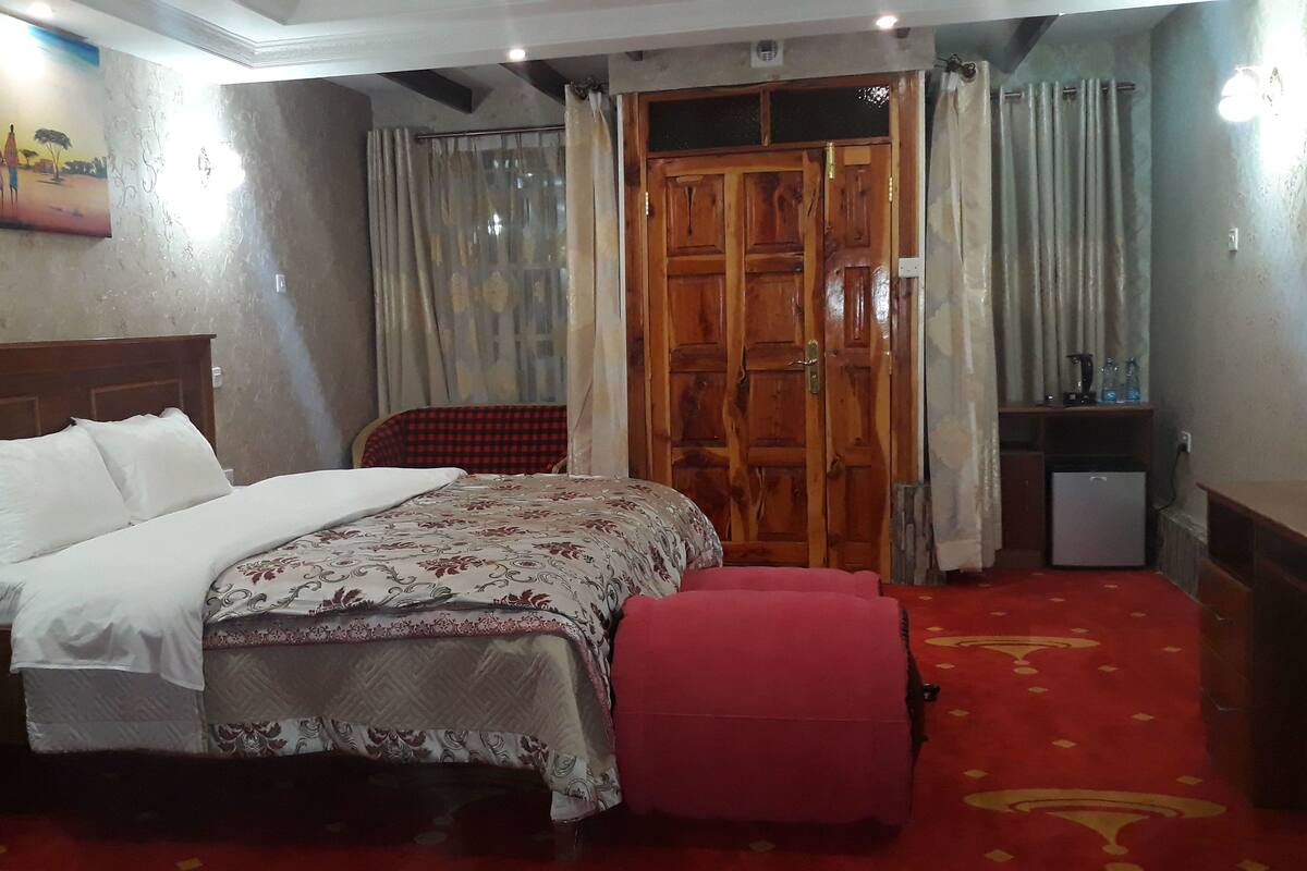 Quarto de hotel com uma cama, um guarda-roupa de madeira, uma mesa pequena com uma luminária e um quadro na parede.
