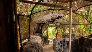 Un paseo techado con grandes rocas, bancos de madera y una estructura de techo de malla.