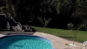 Una piscina con agua azul cristalina, rodeada de baldosas de piedra y una zona ajardinada con árboles.