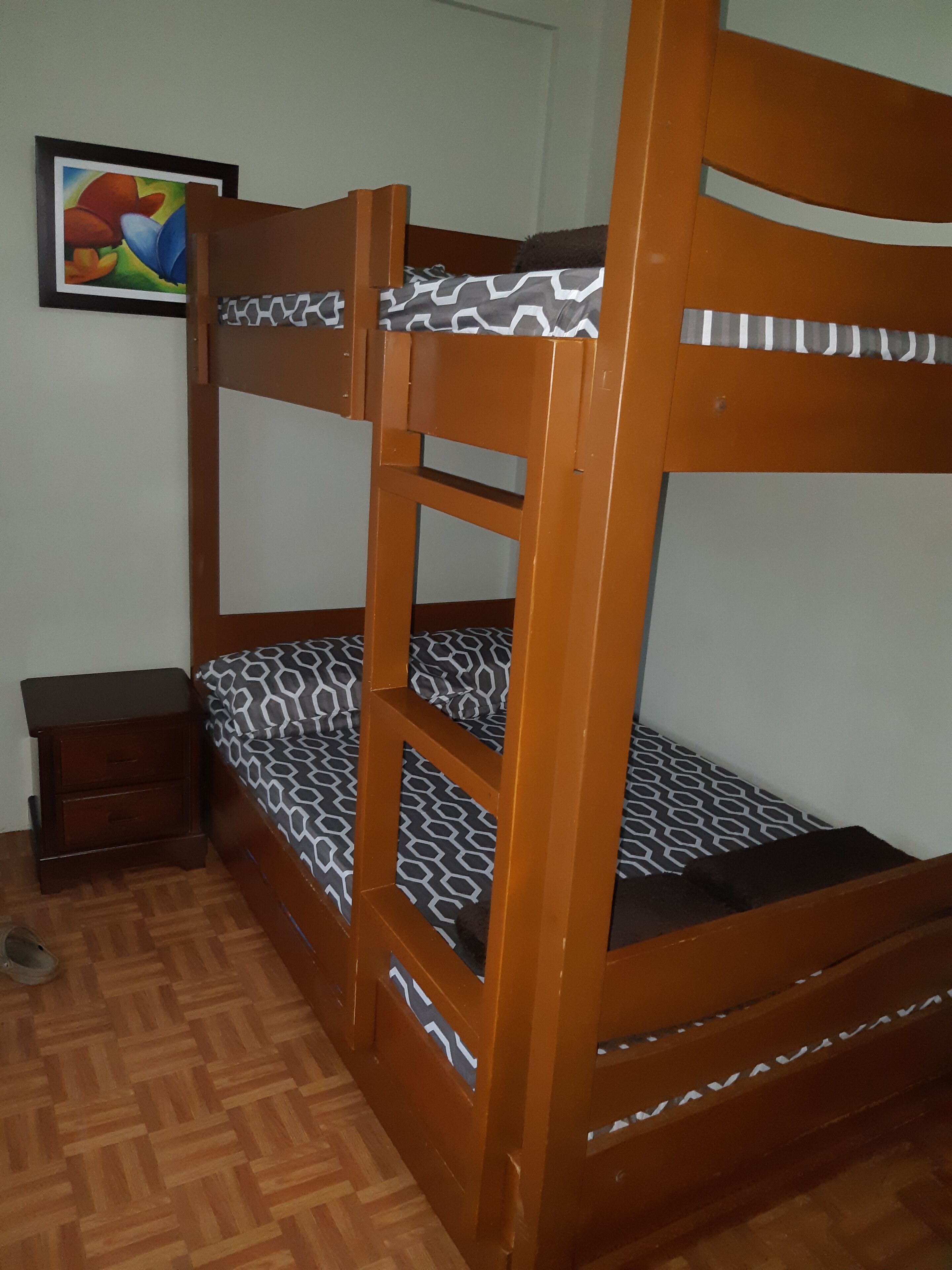 Een stapelbed met een bekleed matras, een houten nachtkastje en een lijst met een foto aan de muur.