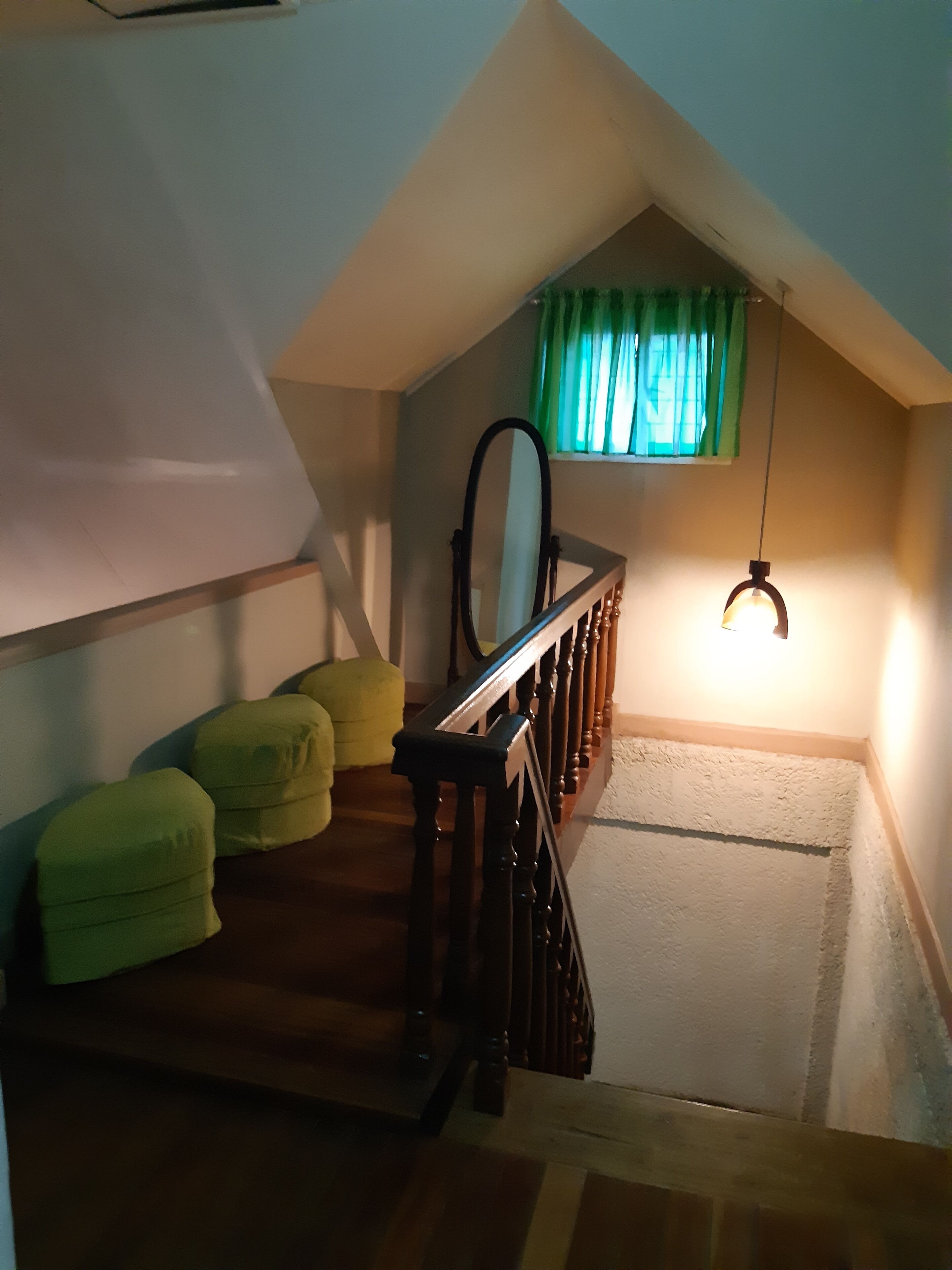 Un escalier menant à une chambre à coucher située en mezzanine, avec une rambarde en bois, un coussin vert et un luminaire suspendu.