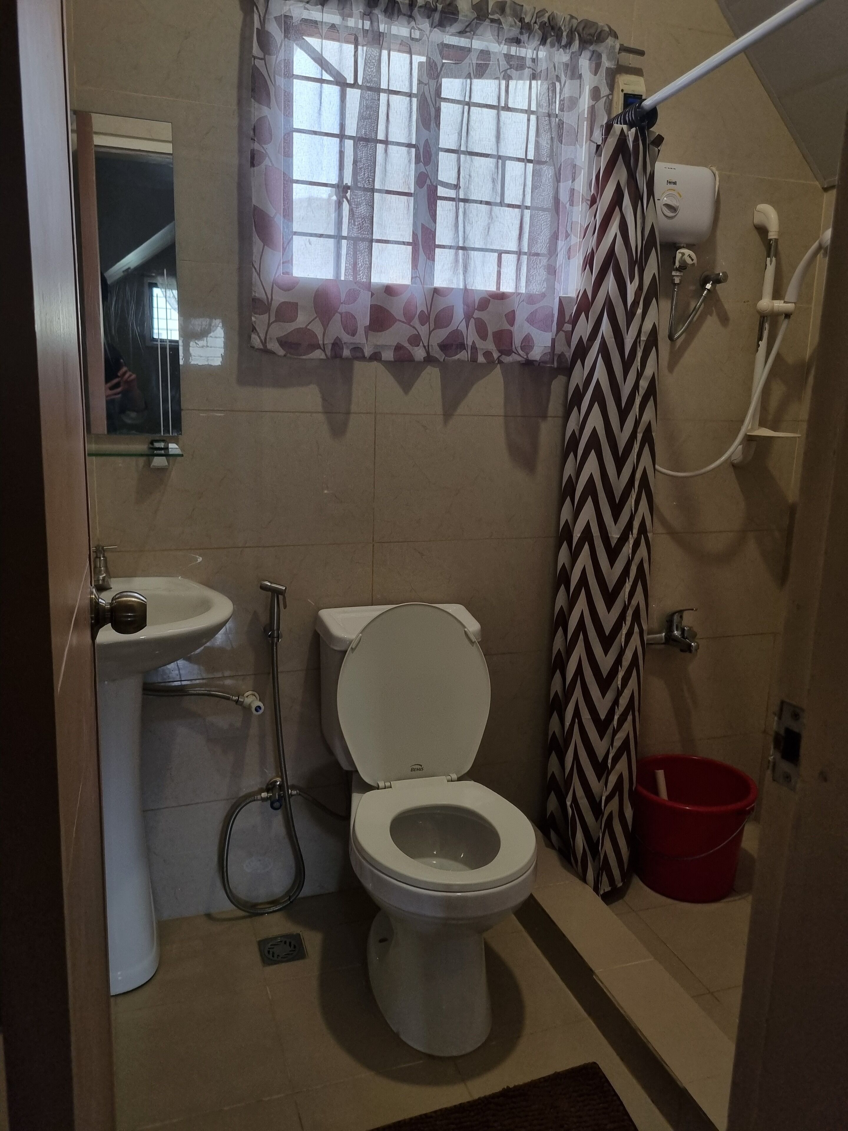 A bathroom with a toilet, sink, shower, and a window with a patterned curtain.