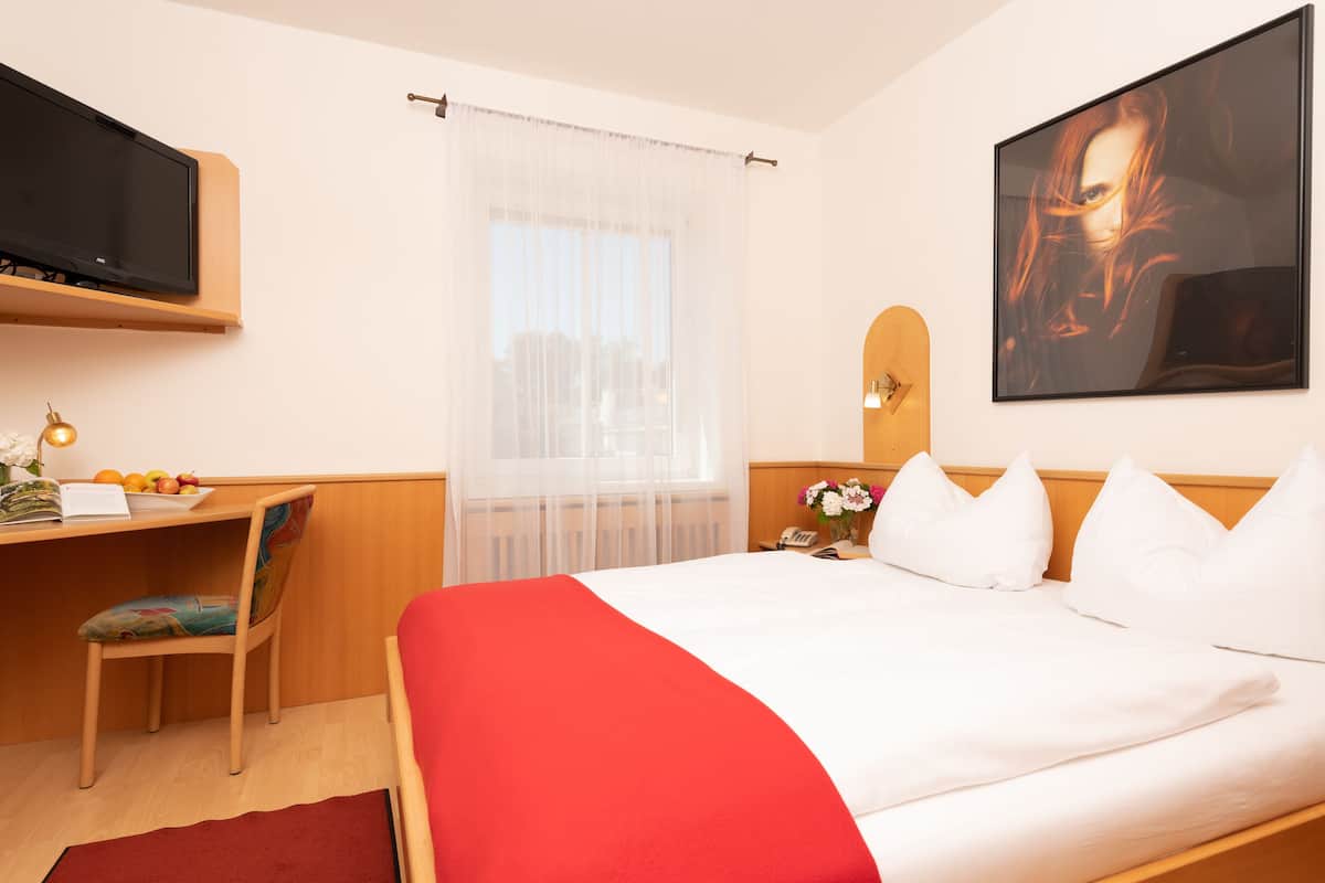 Une chambre d’hôtel avec un lit, un bureau et une chaise, une télévision, une fenêtre avec des rideaux légers et un tableau au mur.