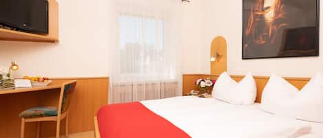 Une chambre d’hôtel avec un lit, un bureau et une chaise, une télévision, une fenêtre avec des rideaux légers et un tableau au mur.