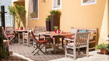 An outdoor patio with wooden tables and chairs, potted plants, and a brick floor.