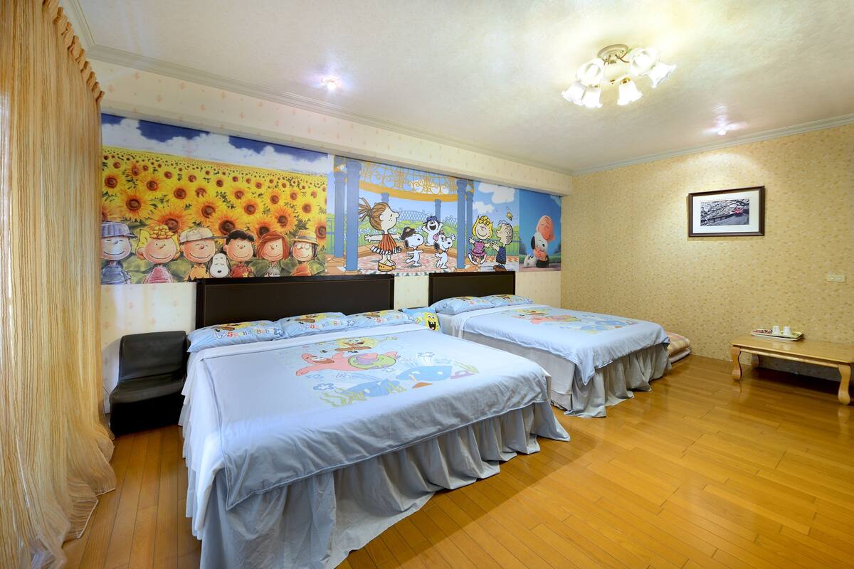 Habitación de hotel con tres camas, un mural de personajes de dibujos animados, suelo de madera y una pequeña mesa con una lámpara.